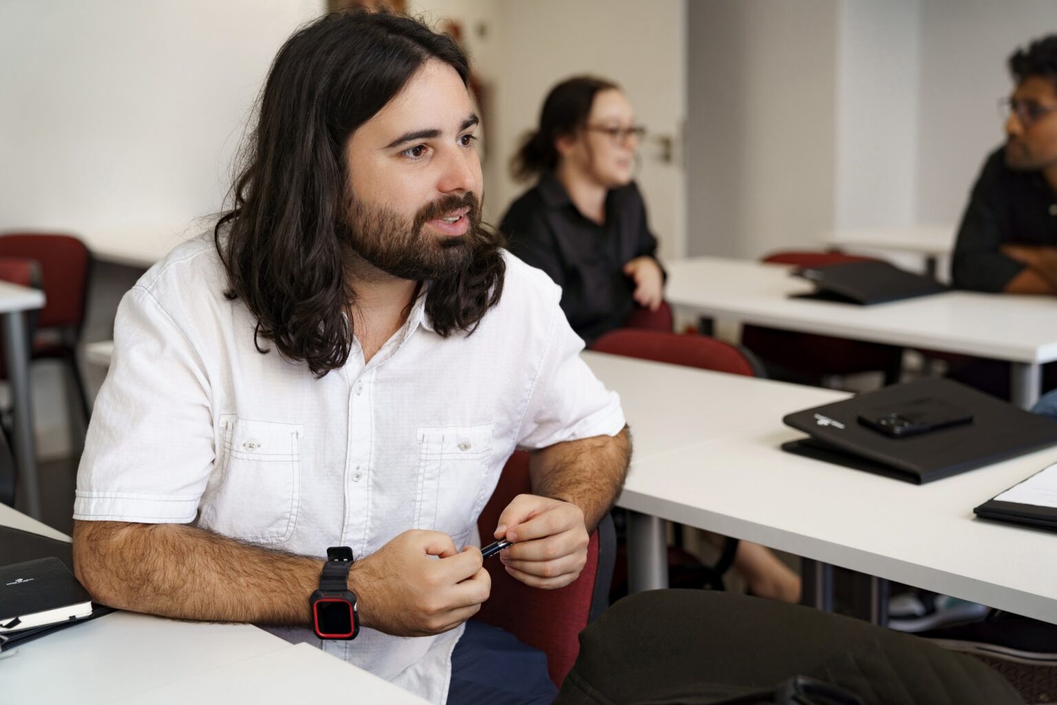 Student with long hair and a beard speaking during a class discussion at GBSB Global, classmates at their desks behind him